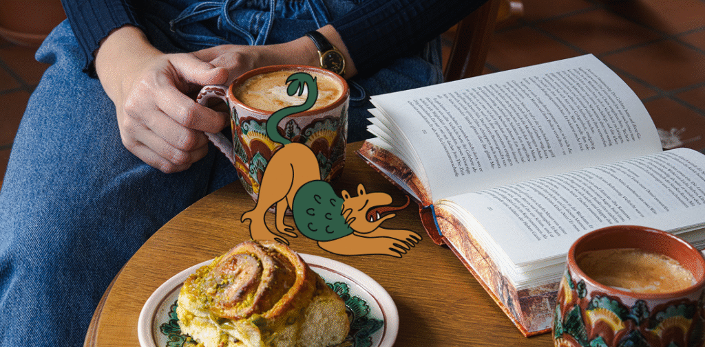 Someone sitting at a table with an open book, coffee in a Kosiv ceramic cup, a delcious pastry, with a superimposed illustration of Cozy Leo stretching on the table